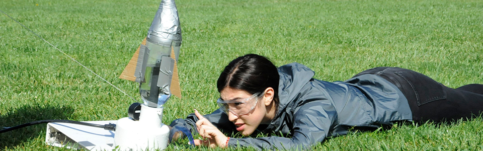 A student lies in the grass, setting up a bottle rocket